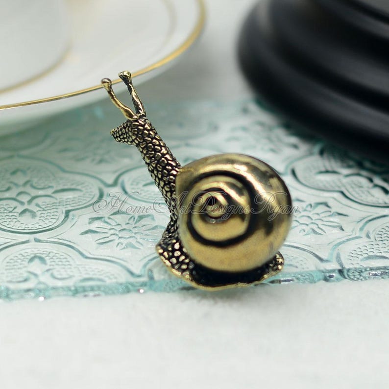 Puede incluir: Una figurita de caracol de metal dorado con una concha detallada y un cuerpo texturizado. El caracol est&aacute; sentado sobre una superficie de vidrio azul claro.