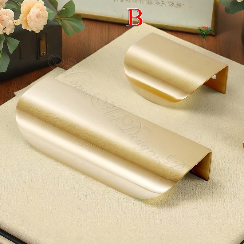 K&ouml;nnte beinhalten: Zwei goldfarbene, gebogene Schrankgriffe. Die Griffe haben ein schlankes, modernes Design mit glatter, metallischer Oberfl&auml;che. Die Griffe sind aus Metall gefertigt und zur Montage an einer Schrankt&uuml;r oder Schublade vorgesehen.