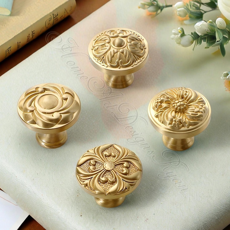 May include: Four antique brass cabinet knobs with detailed floral patterns. Each knob has a unique embossed design, featuring swirling leaves and flower motifs. The knobs are presented on a light surface, highlighting their intricate craftsmanship. The knobs are made of brass.