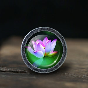 K&ouml;nnte beinhalten: Ein runder, silberfarbener Schrank-Knopf mit einem Glaseinsatz, der eine rosa und violette Lotusblume auf gr&uuml;nem Hintergrund zeigt.