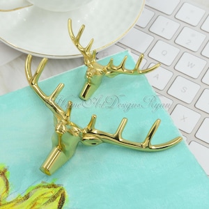 Puede incluir: Dos ganchos de cabeza de ciervo de color dorado con cuernos. Los ganchos están sobre una superficie turquesa.