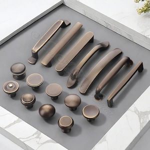 K&ouml;nnte beinhalten: Eine Auswahl an bronzefarbenen Schrankbeschl&auml;gen, darunter verschiedene Griffstile und runde Kn&ouml;pfe. Die Griffe sind gebogen und gerade, w&auml;hrend die Kn&ouml;pfe kuppelf&ouml;rmig sind. Die Beschl&auml;ge sind auf einer grauen Oberfl&auml;che in einem wei&szlig; gerahmten Tablett angeordnet.