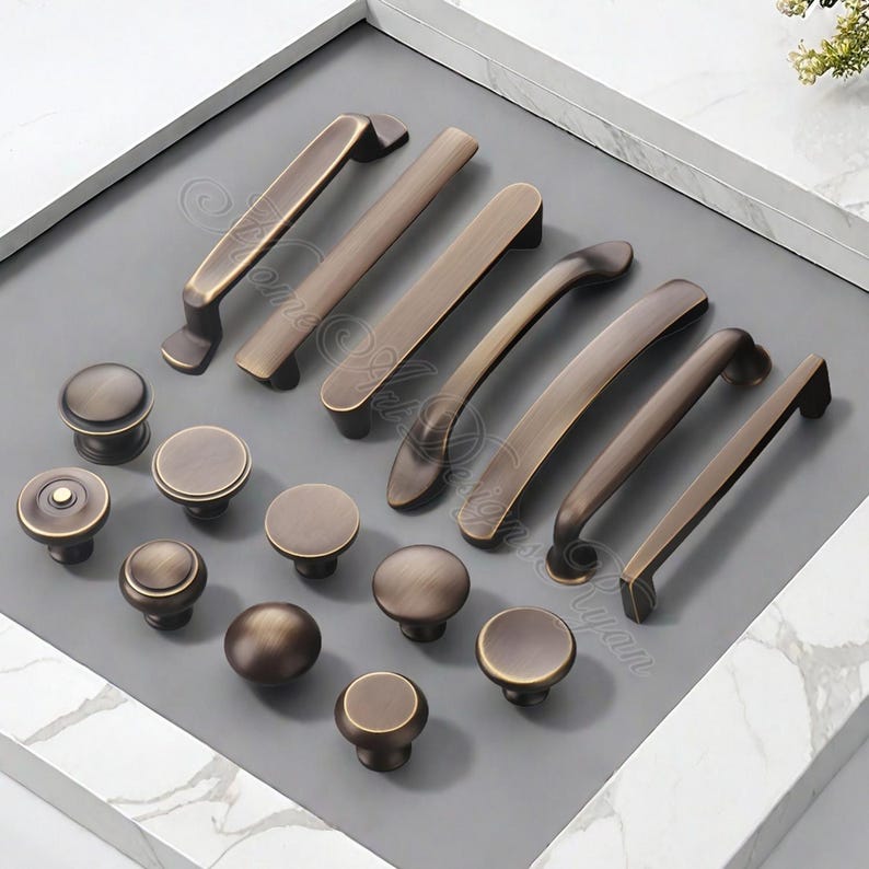 K&ouml;nnte beinhalten: Eine Auswahl an bronzefarbenen Schrankbeschl&auml;gen, darunter verschiedene Griffstile und runde Kn&ouml;pfe. Die Griffe sind gebogen und gerade, w&auml;hrend die Kn&ouml;pfe kuppelf&ouml;rmig sind. Die Beschl&auml;ge sind auf einer grauen Oberfl&auml;che in einem wei&szlig; gerahmten Tablett angeordnet.