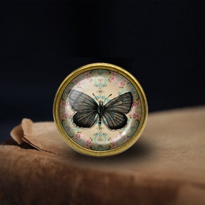 May include: A gold-toned round knob with a glass top featuring a black butterfly on a floral pattern background.