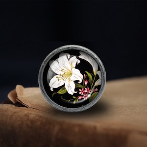 Peut inclure: Un bouton de porte rond en argent avec un fond noir et une fleur de lys blanche avec des fleurs roses en arrière-plan.