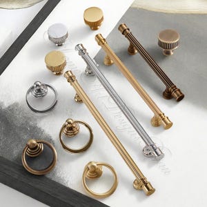 Puede incluir: Una colecci&oacute;n de herrajes para armarios, que incluye pomos y tiradores, en varios acabados met&aacute;licos. La selecci&oacute;n presenta pomos redondos, tiradores de anillo y tiradores de barra en tonos dorados, plateados y bronce. El hardware se muestra sobre una superficie blanca.
