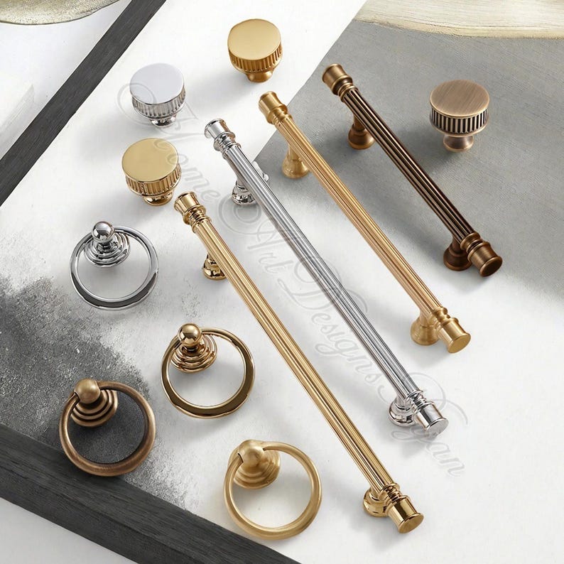 Puede incluir: Una colecci&oacute;n de herrajes para armarios, que incluye pomos y tiradores, en varios acabados met&aacute;licos. La selecci&oacute;n presenta pomos redondos, tiradores de anillo y tiradores de barra en tonos dorados, plateados y bronce. El hardware se muestra sobre una superficie blanca.