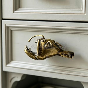 K&ouml;nnte beinhalten: Ein bronzefarbener Schrankgriff in Form eines Anglerfisches. Der Griff weist detaillierte Schuppen, Z&auml;hne und einen K&ouml;der auf. Der Griff ist an einer hellgrauen Schublade befestigt.