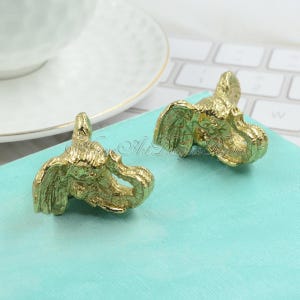 May include: Two gold-coloured elephant head drawer pulls. The elephant heads are facing the right side of the image.
