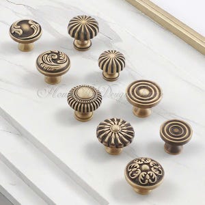 Pu&ograve; includere: Una collezione di pomelli per mobili in ottone antico con vari disegni decorativi. I pomelli presentano motivi floreali, a spirale e a coste. I pomelli sono disposti su una superficie bianca.