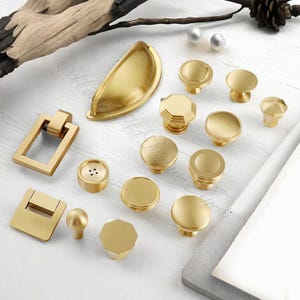 May include: A collection of gold-toned cabinet hardware, including various knobs, pulls, and a handle. The hardware features different shapes, such as round, octagonal, and rectangular designs. The items are arranged on a white surface, showcasing their metallic finish.