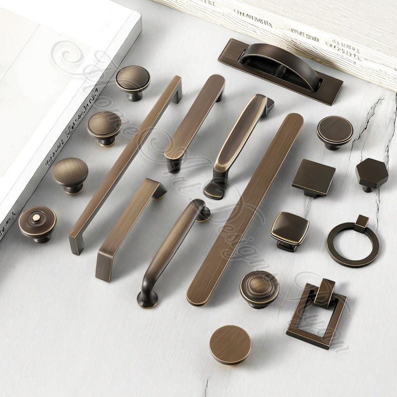 K&ouml;nnte beinhalten: Eine Auswahl an bronzefarbener Schrankbeschl&auml;gen, darunter Kn&ouml;pfe, Griffe und Ziehgriffe. Die Kollektion umfasst verschiedene Formen und Gr&ouml;&szlig;en, von runden Kn&ouml;pfen bis hin zu rechteckigen Ziehgriffen und Griffen, alle mit einem Vintage-Look.