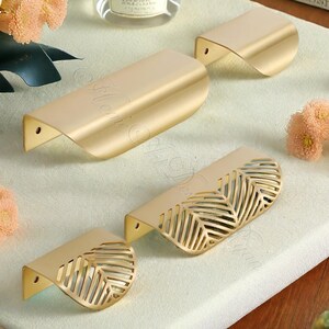 K&ouml;nnte beinhalten: Eine Sammlung goldfarbener Schrankgriffe. Einige Griffe haben ein durchgehendes, gebogenes Design, w&auml;hrend andere ein blattartiges Ausschnittmuster aufweisen. Die Hardware ist auf einer hellen Oberfl&auml;che angeordnet, was auf ein Wohndekor-Thema hindeutet.