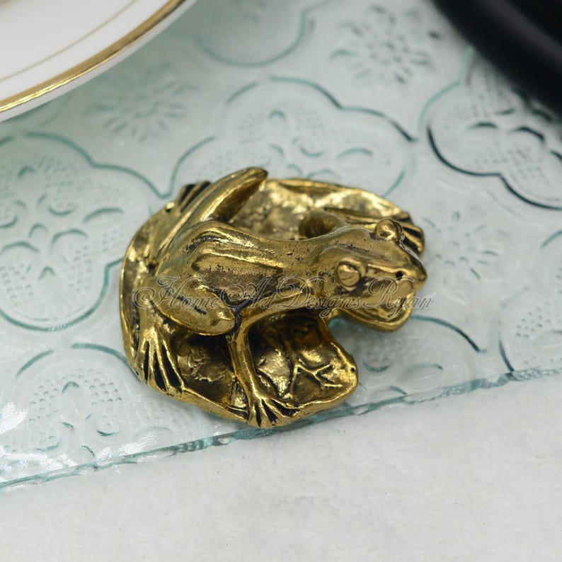 May include: A gold-toned frog figurine sitting on a leaf. The frog is detailed with textured skin and realistic features. The figurine is a decorative piece for a home or office.