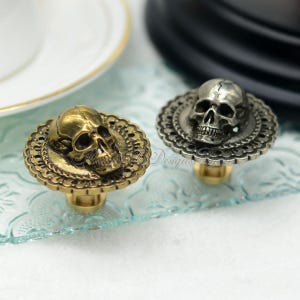 May include: Two cabinet knobs featuring skull designs. One is gold-toned, the other silver. Each skull sits on an ornate, circular base. The knobs have a vintage, gothic aesthetic, suitable for furniture or decor. The knobs are likely made of metal.