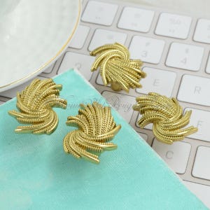 Può includere: Quattro pomelli decorativi dorati con un design a spirale e strutturato. I pomelli sono disposti su una superficie azzurra, con una tastiera bianca sullo sfondo. I pomelli hanno un aspetto dettagliato e ornato.