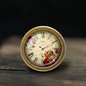 Puede incluir: Una esfera de reloj de color dorado con números romanos y un diseño floral. La esfera del reloj está rodeada por un marco de metal dorado.