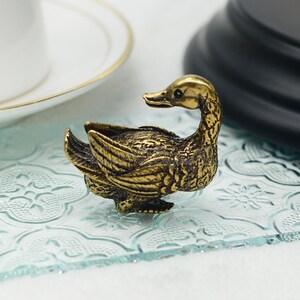 Op de afbeelding: Een klein, goudkleurig metalen eendje met ingewikkelde details. Het eendje zit op een lichtblauw, gepatroneerd oppervlak.