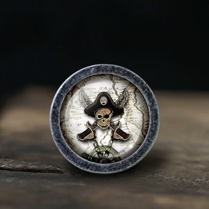May include: A round, silver-colored knob with a pirate skull and crossbones design on a vintage map background. The skull is wearing a black pirate hat.