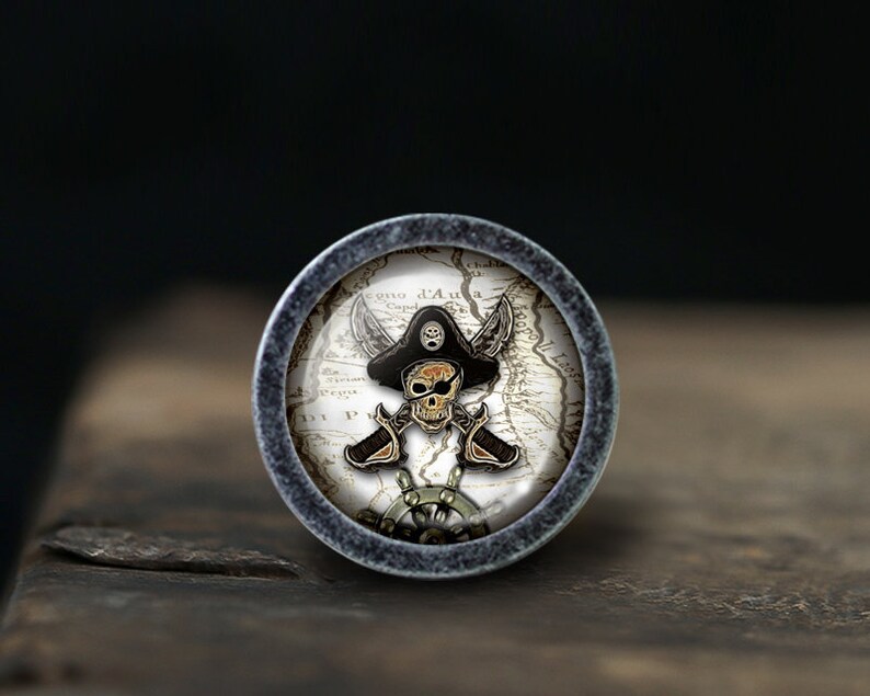 May include: A round, silver-colored knob with a pirate skull and crossbones design on a vintage map background. The skull is wearing a black pirate hat.