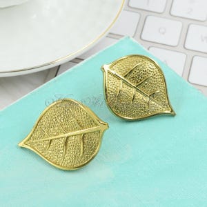 Puede incluir: Un par de pendientes de hoja de oro. Los pendientes son texturizados y tienen un diseño ligeramente elevado.