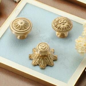 May include: Three brass cabinet knobs with floral designs. The knobs are in various shapes, including a round, a domed, and a starburst design. The knobs are displayed on a light blue surface within a gold-framed tray.