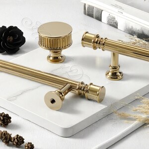 Puede incluir: Juego de herrajes dorados para armarios, que incluye un pomo y dos estilos diferentes de tiradores. Los herrajes presentan un dise&ntilde;o acanalado y un acabado pulido. El juego se muestra sobre una superficie blanca con un patr&oacute;n similar al m&aacute;rmol.