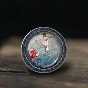 May include: A round, silver-colored knob with a glass insert featuring a watercolor illustration of a girl in a pink dress standing on a blue bubble. The background is a soft blue with a pink flower.