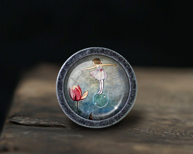 May include: A round, silver-colored knob with a glass insert featuring a watercolor illustration of a girl in a pink dress standing on a blue bubble. The background is a soft blue with a pink flower.
