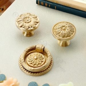 May include: Three antique brass cabinet hardware pieces. Two are round knobs with floral designs, and the third is a ring pull with a similar floral motif. The hardware is displayed on a light-coloured surface, suggesting a vintage or classic aesthetic.