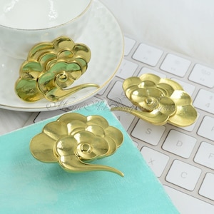 Może przedstawiać: Trzy złote metalowe uchwyty do szuflad w kształcie stylizowanych chmur. Uchwyty mają spiralny wzór i wypukłe centrum.