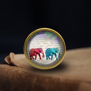 May include: A gold-toned round glass cabochon featuring a vintage-style image of two elephants. The elephants are pink and blue with floral patterns. The background is white paper with cursive writing.