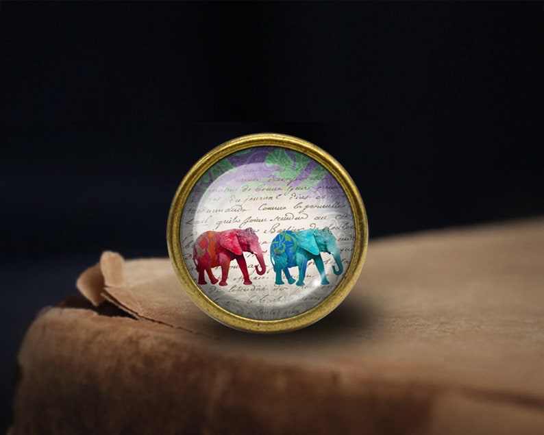 May include: A gold-toned round glass cabochon featuring a vintage-style image of two elephants. The elephants are pink and blue with floral patterns. The background is white paper with cursive writing.