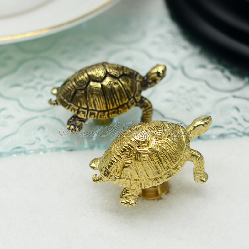 May include: Two gold-toned turtle-shaped drawer pulls. The turtles have detailed shell patterns and are facing different directions.