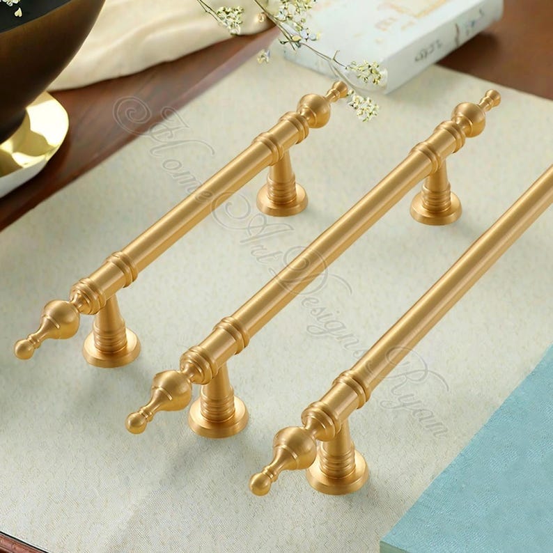 Puede incluir: Tres tiradores dorados para armarios con un dise&ntilde;o cl&aacute;sico. Cada tirador presenta una barra larga y recta con remates decorativos en cada extremo y un soporte central. Los tiradores son de metal y tienen un acabado liso y mate.