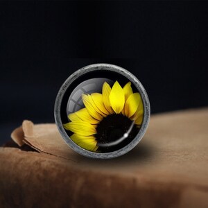 Op de afbeelding: Een zilveren knop met een glazen inzetstuk met een gele zonnebloem.