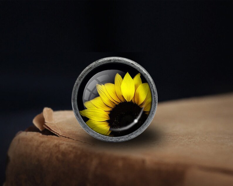 Op de afbeelding: Een zilveren knop met een glazen inzetstuk met een gele zonnebloem.