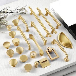 May include: A collection of gold-toned cabinet hardware, including knobs, pulls, and handles, arranged on a white surface. The hardware features various designs, from simple round knobs to ornate pulls, all with a polished finish. The assortment offers options for kitchen or furniture upgrades.