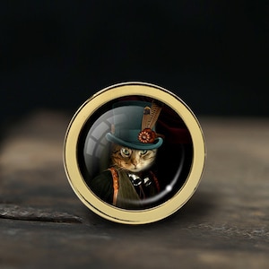 Könnte beinhalten: Ein runder Anhänger aus goldfarbenem Metall mit einer Glaskuppel, die ein Kätzchen zeigt, das einen grünen Zylinder mit einer Feder und einer roten Blume trägt. Die Katze hat grüne Augen und ein braunes und weißes gestreiftes Fell.