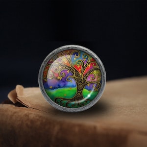 Peut inclure: Un pendentif rond argenté avec un motif d'arbre de vie coloré. L'arbre a un tronc brun et des branches avec des motifs en spirale dans des tons de rose, violet, bleu, vert et jaune. L'arrière-plan est un dégradé de bleu, violet et vert.