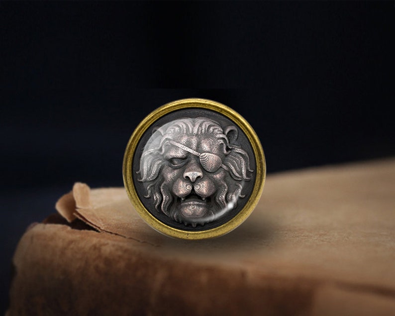 Puede incluir: Un bot&oacute;n circular con un borde dorado y un dise&ntilde;o detallado de cabeza de le&oacute;n de plata. El le&oacute;n tiene un parche negro sobre su ojo derecho.