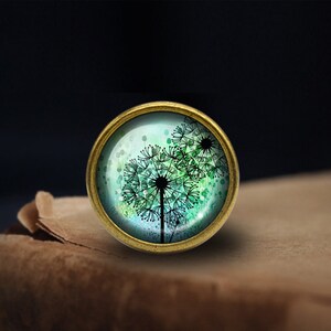 May include: A round, brass-coloured pendant with a glass dome featuring a watercolour-style image of a dandelion with green and blue hues.
