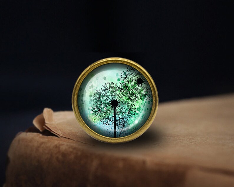 May include: A round, brass-coloured pendant with a glass dome featuring a watercolour-style image of a dandelion with green and blue hues.