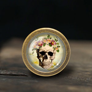 K&ouml;nnte beinhalten: Ein Messingknopf mit einem Glaseinsatz, der einen Sch&auml;del mit einer Blumenkrone zeigt. Der Sch&auml;del ist wei&szlig; mit schwarzen Augenh&ouml;hlen und einem schwarzen Mund. Die Blumen sind rosa, wei&szlig; und gelb.