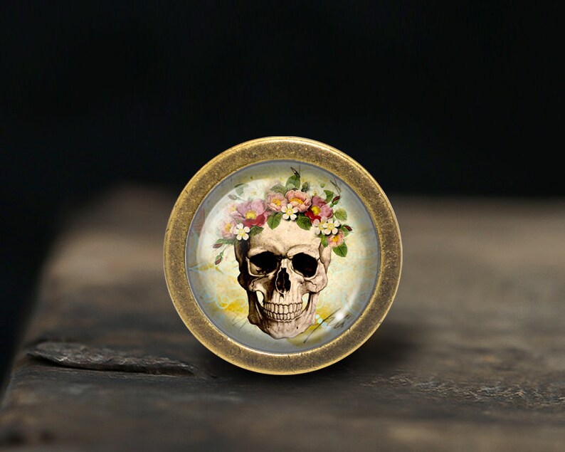 K&ouml;nnte beinhalten: Ein Messingknopf mit einem Glaseinsatz, der einen Sch&auml;del mit einer Blumenkrone zeigt. Der Sch&auml;del ist wei&szlig; mit schwarzen Augenh&ouml;hlen und einem schwarzen Mund. Die Blumen sind rosa, wei&szlig; und gelb.