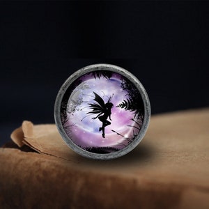 May include: A silver-toned metal charm with a glass cabochon featuring a silhouette of a fairy flying in front of a purple and pink moon with stars. The fairy has wings and is holding a flower.
