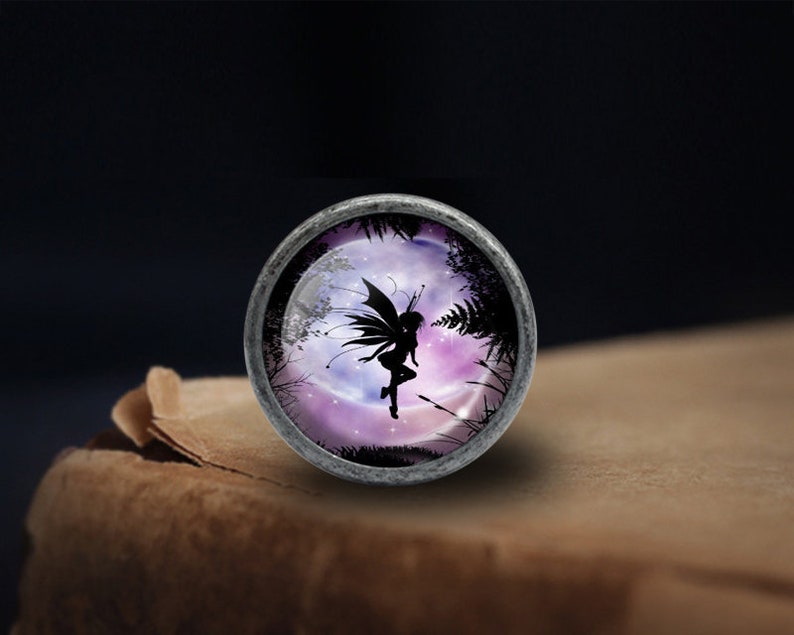 May include: A silver-toned metal charm with a glass cabochon featuring a silhouette of a fairy flying in front of a purple and pink moon with stars. The fairy has wings and is holding a flower.