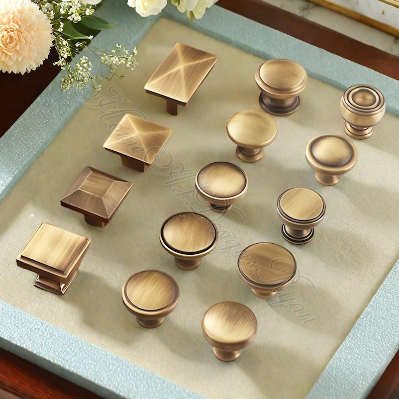 May include: An assortment of antique brass cabinet knobs in diverse shapes, including square, rectangular, and round designs. The knobs have a warm, aged finish and are displayed on a light-coloured surface.