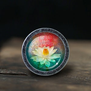 May include: A silver-toned ring with a round glass cabochon featuring a colourful image of a white water lily with a yellow centre. The background is a gradient of red, orange, yellow, green, and blue.