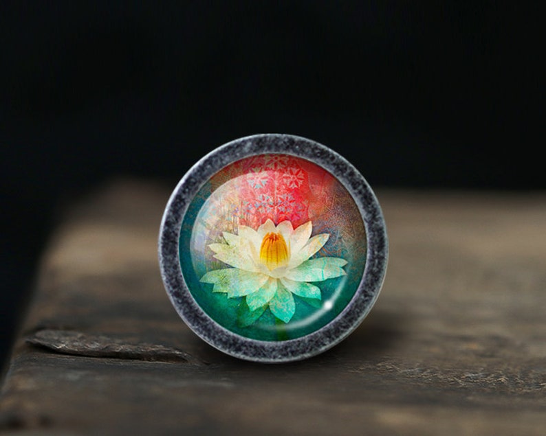 May include: A silver-toned ring with a round glass cabochon featuring a colorful image of a white water lily with a yellow center. The background is a gradient of red, orange, yellow, green, and blue.